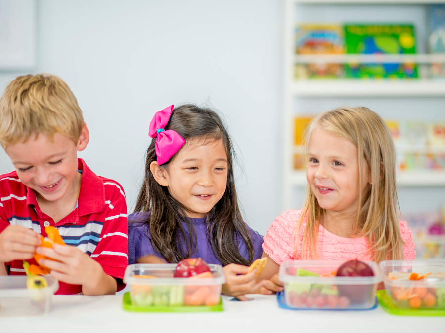A multi-ehthnic group of elementary age children are sitting together at a table eating their healthy snacks full of apples, carrots, and celery.